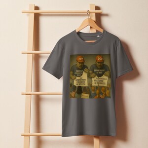 Può includere: Una maglietta grigia con una grafica in stile vintage. La grafica raffigura due figure con cappelli con le etichette "FCC" e "DOD", che tengono cartelli sulla libertà di parola e di stampa. Il testo sulla maglietta include "Fascist Censorship Cult" e "Department of War".