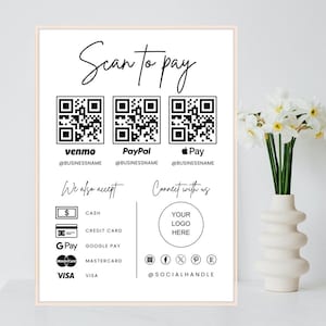 May include: A framed white sign with the text "Scan to pay" and QR codes for Venmo, PayPal, and Apple Pay. It also lists accepted payment methods: cash, credit card, Google Pay, Mastercard, and Visa. A vase of white flowers is on the right.