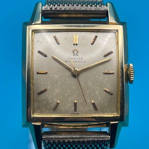 May include: A vintage Omega Automatic wristwatch with a gold-toned rectangular case and a mesh band. The watch face is a light color with gold hands and markers. The watch face has the Omega logo and the words "OMEGA AUTOMATIC" and "SWISS".
