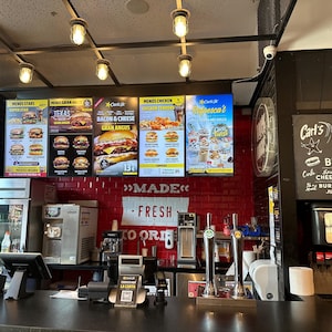 May include: Interior shot of a Carl's Jr. restaurant with illuminated menu boards displaying burger options. A counter with ordering equipment and drink dispensers is in the foreground. The sign says "Made Fresh To Order".