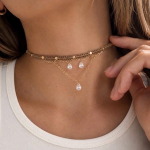 May include: A beige woven cord choker necklace featuring small gold-toned metal beads and two delicate gold-toned chains. The chains are adorned with three clear, teardrop-shaped crystals that hang in a layered arrangement around the neck.