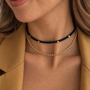 May include: A layered necklace set consisting of a dark blue cord choker featuring small gold-toned metallic beads and a separate gold-toned chain necklace with round interlocking links, worn together around the neck.