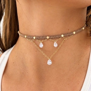 May include: A layered choker necklace with a tan macrame band and gold-colored accents. Three gold chains hang down, each with a clear teardrop-shaped pendant. The necklace is worn around the neck.