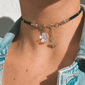Peut inclure: Un collier ras-du-cou en cordon bleu foncé avec des éléments métalliques dorés, une breloque en forme de fleur à six pétales ornée de pierres transparentes à facettes et une petite breloque abeille dorée suspendue à une fine chaînette.