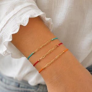 May include: Three bracelets featuring gold-toned paperclip chains connected to colorful nylon cords. The top bracelet has a turquoise cord, the middle has a red cord, and the bottom has an orange cord. Each chain segment is accented with small gold beads.