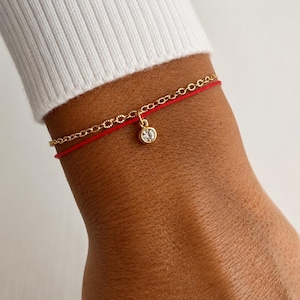 May include: A double-strand bracelet featuring one thin gold chain and one smooth red cord. A single round, clear, bezel-set gemstone charm hangs from the center of the gold chain, resting against the red cord. The bracelet is worn on a wrist.