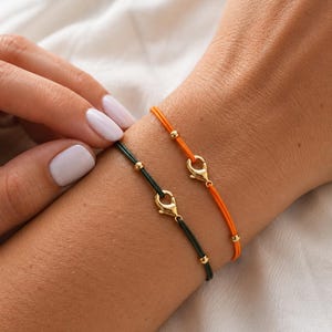 May include: Two cord bracelets, one in dark green and one in bright orange, each featuring a gold-toned lobster claw clasp and small gold-toned metal beads spaced along the cord. The bracelets are worn together on a wrist, showcasing the contrast.