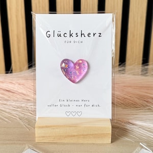 Könnte beinhalten: Ein herzförmiger Ansteckpin mit rosa und lila Glitzerdesign, präsentiert auf einer weißen Karte mit deutschem Text. Die Karte ist auf einem kleinen Holzständer platziert. Weitere Karten mit Herz-Pins sind sichtbar.