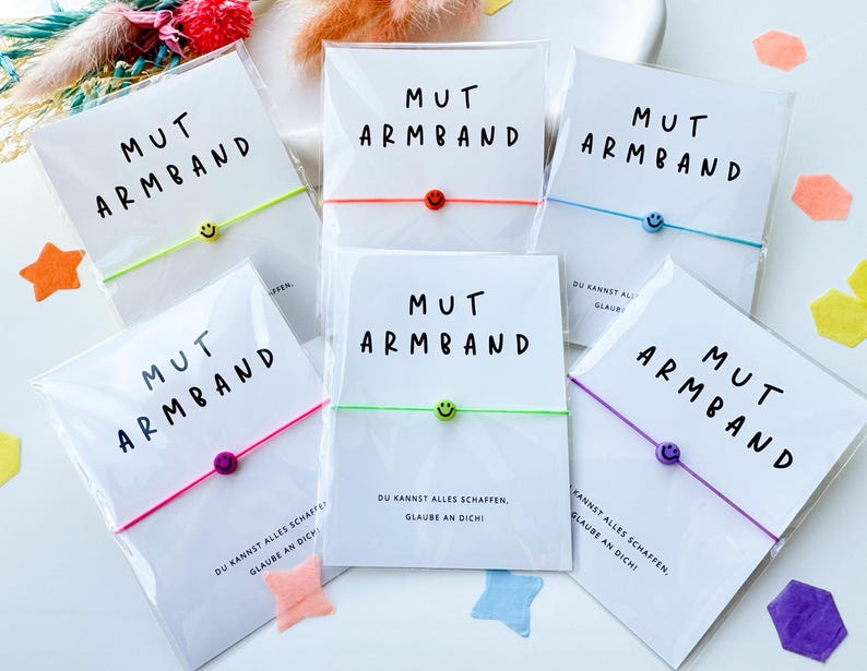 May include: A collection of bracelets with the text "MUT ARMBAND" on white cards. Each bracelet has a different colored cord and a smiley face charm. The cards also have the text "DU KANNST ALLES SCHAFFEN, GLAUBE AN DICH!"