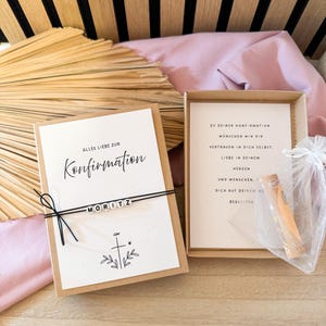 May include: A gift box with a card that reads "Alles Liebe zur Konfirmation" and the name "MORITZ". The box also contains a card with text and a small item in a clear bag. The background includes wooden slats and pink fabric.