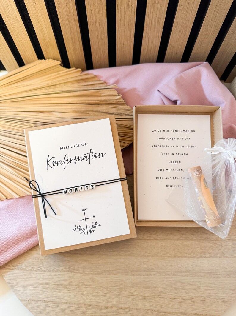 May include: A gift box with a card that reads "Alles Liebe zur Konfirmation" and the name "MORITZ". The box also contains a card with text and a small item in a clear bag. The background includes wooden slats and pink fabric.