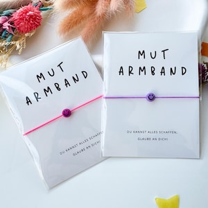 May include: Two bracelets on white cards with the text "MUT ARMBAND" and a smiley face bead. One bracelet is pink, the other is purple. The cards also have the text "DU KANNST ALLES SCHAFFEN, GLAUBE AN DICH!"