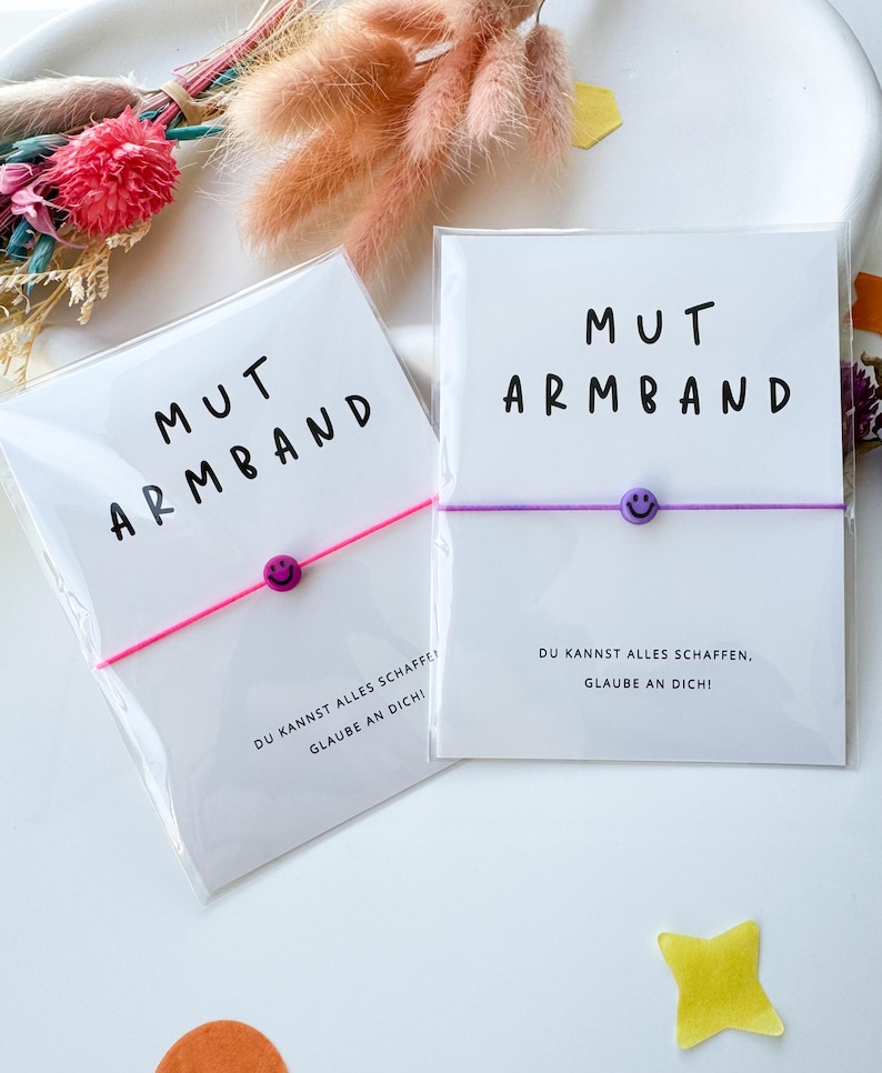 May include: Two bracelets on white cards with the text "MUT ARMBAND" and a smiley face bead. One bracelet is pink, the other is purple. The cards also have the text "DU KANNST ALLES SCHAFFEN, GLAUBE AN DICH!"