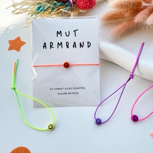 May include: A white card with the words "MUT ARMBAND" and a neon orange bracelet with a smiley face bead. The card also has the text "DU KANNST ALLES SCHAFFEN. GLAUBE AN DICH!" Several other bracelets in neon colors with smiley face beads are also visible.