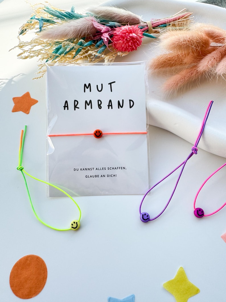 May include: A white card with the words "MUT ARMBAND" and a neon orange bracelet with a smiley face bead. The card also has the text "DU KANNST ALLES SCHAFFEN. GLAUBE AN DICH!" Several other bracelets in neon colors with smiley face beads are also visible.