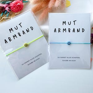 May include: Two bracelets with the text "MUT ARMBAND" and a smiley face charm. One bracelet has a neon green band, the other a light blue band. The cards read "DU KANNST ALLES SCHAFFEN, GLAUBE AN DICH!"
