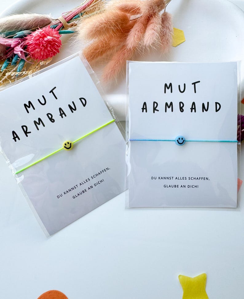 May include: Two bracelets with the text "MUT ARMBAND" and a smiley face charm. One bracelet has a neon green band, the other a light blue band. The cards read "DU KANNST ALLES SCHAFFEN, GLAUBE AN DICH!"