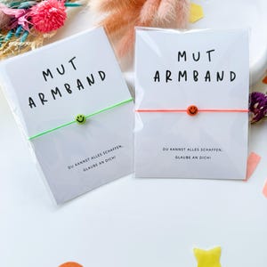 May include: Two bracelets with the text "MUT ARMBAND" on the packaging. One bracelet has a green cord and a green smiley face bead, the other has an orange cord and an orange smiley face bead. The packaging also includes the text "DU KANNST ALLES SCHAFFEN, GLAUBE AN DICH!"