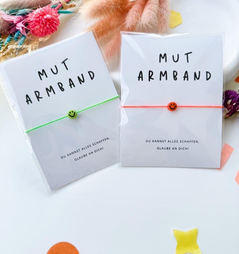 May include: Two bracelets with the text "MUT ARMBAND" on the packaging. One bracelet has a green cord and a green smiley face bead, the other has an orange cord and an orange smiley face bead. The packaging also includes the text "DU KANNST ALLES SCHAFFEN, GLAUBE AN DICH!"