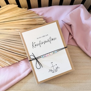 May include: A greeting card with the words "Alles Liebe zur Konfirmation" and the name "Bastian" spelled out with colorful beads on a black string. The card is on a wooden surface with a pink cloth and a dried palm leaf.