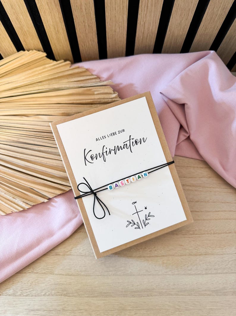 May include: A greeting card with the words "Alles Liebe zur Konfirmation" and the name "Bastian" spelled out with colorful beads on a black string. The card is on a wooden surface with a pink cloth and a dried palm leaf.