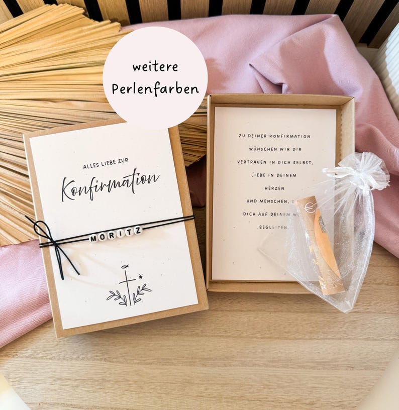 May include: A gift box with a personalized card that reads "Alles Liebe zur Konfirmation" and the name "MORITZ". The box also contains a small gift in a sheer bag and a card with text. The background includes a pink fabric and wooden elements.