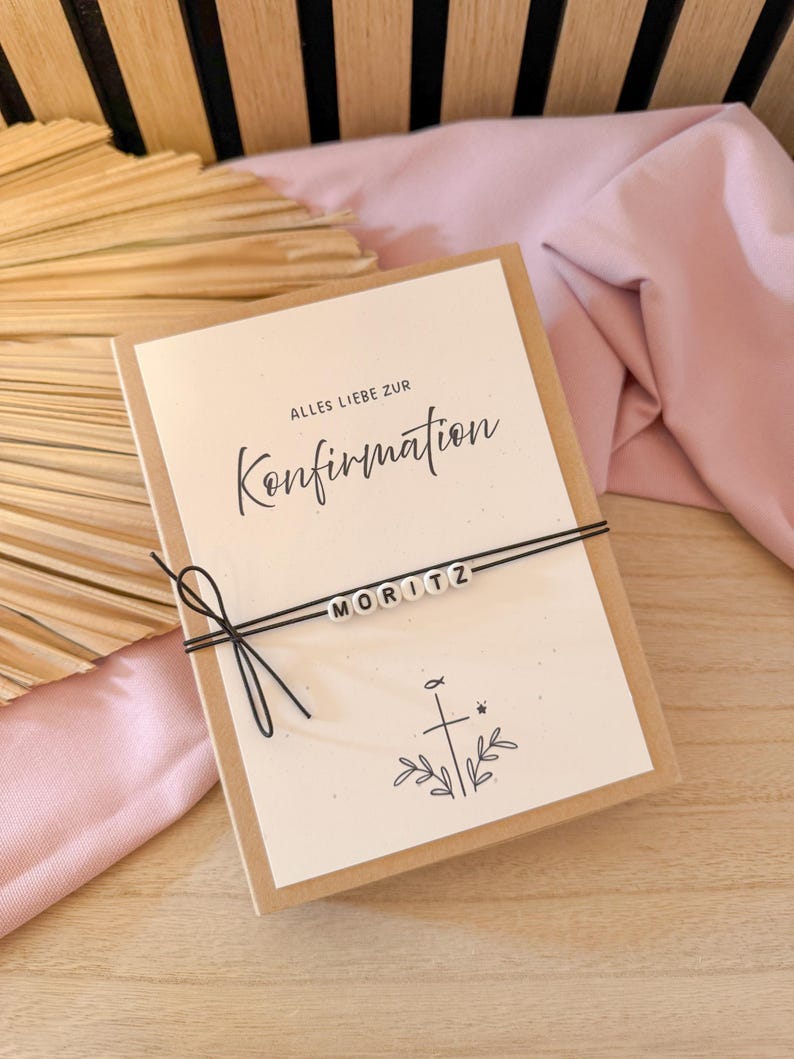 May include: A greeting card with the words "Alles Liebe zur Konfirmation" and the name "MORITZ" spelled out in white beads. The card is tied with black string and features a cross and leaf design.