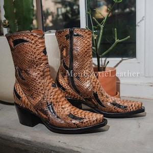 May include: A pair of brown and black leather cowboy boots with a snakeskin pattern. The boots have a pointed toe, a mid-height heel, and a zipper closure. The boots are displayed on a ledge near a window, with a cactus in a white pot.