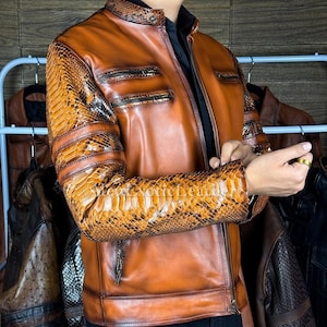 Op de afbeelding: Een bruine leren jas met slangenprint op de mouwen en schouders. De jas heeft een ritssluiting, meerdere zakken en een opstaande kraag. Andere leren jassen zijn zichtbaar op de achtergrond.