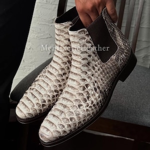 Peut inclure: Une paire de bottines Chelsea blanches et grises à motif peau de serpent. Les bottines ont un empiècement latéral élastique marron et une semelle marron foncé. Le texte "Men Exotic Leather" est visible en bas de l'image.