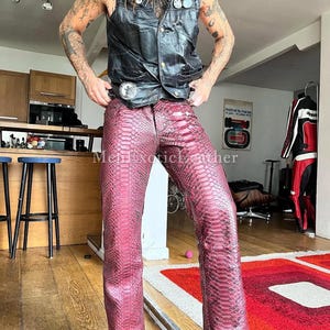 May include: Burgundy snake-skin trousers with a matching leather waistcoat. The trousers feature a detailed scale pattern and a classic straight-leg cut. The waistcoat is black and buttoned. The outfit is paired with brown cowboy boots and a belt with a silver buckle.