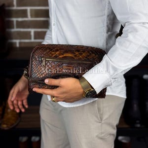 May include: A brown leather travel bag with a snakeskin pattern. The bag features a zip closure and a wrist strap. The text "MenExoticLeather" is visible. The bag is held by a person wearing a white shirt and beige trousers.