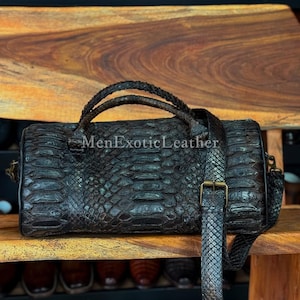Puede incluir: Bolso de cuero cilíndrico marrón oscuro con un patrón texturizado similar al de un reptil. El bolso tiene un asa trenzada y una correa para el hombro desmontable con hebilla metálica. El texto "MenExoticLeather" es visible.