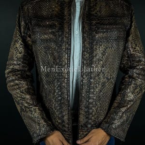 Op de afbeelding: Bruin en zwart leren jack met slangenpatroon, opstaande kraag en zakken met ritssluiting. De jas is open en onthult een lichtblauw overhemd eronder. De tekst "MenExoticLeather" is zichtbaar.