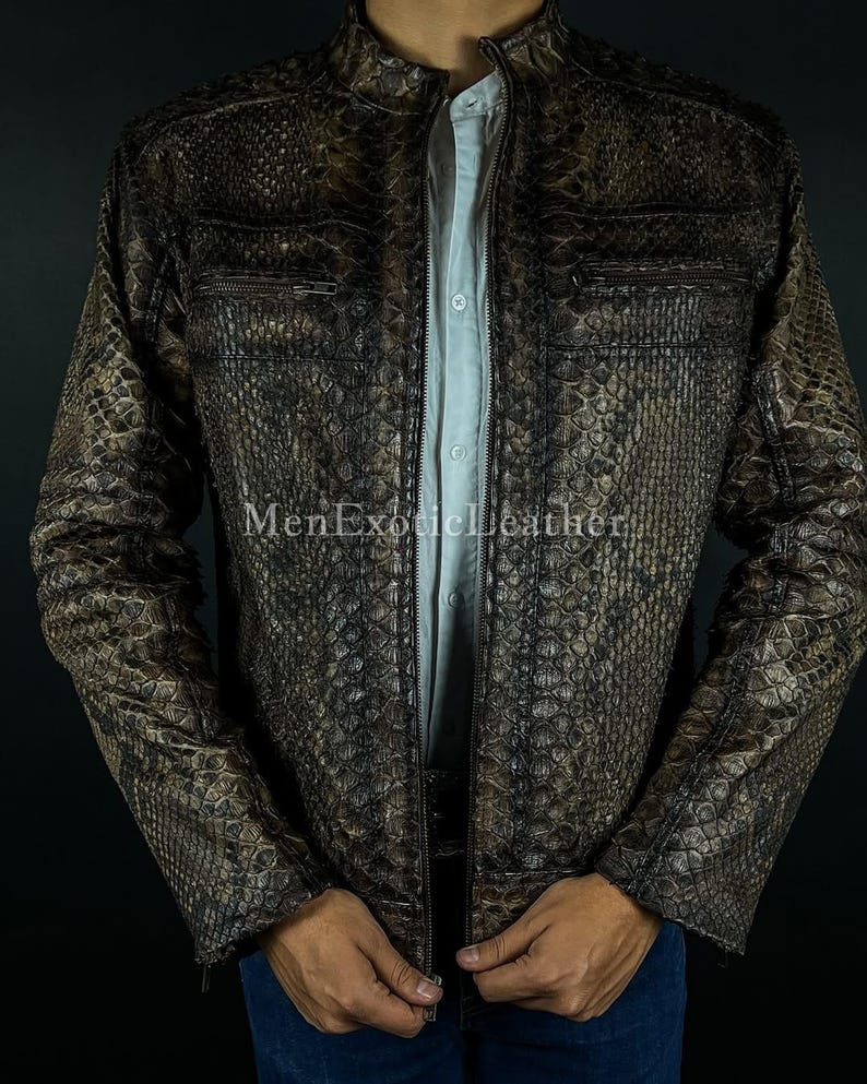 May include: A brown and black snakeskin-patterned leather jacket with a stand-up collar and zippered pockets. The jacket is open, revealing a light blue shirt underneath. The text "MenExoticLeather" is visible on the jacket.