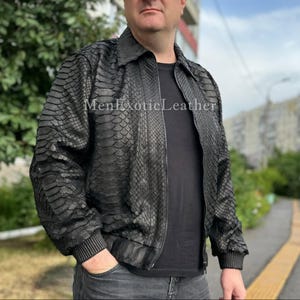 Op de afbeelding: Zwarte leren jas met een textuur van slangenleer. De jas heeft een kraag, een ritssluiting en geribde manchetten en zoom. De tekst "MenExoticLeather" is zichtbaar op de jas.
