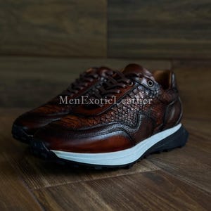 May include: Brown leather trainers with a textured snakeskin pattern. The shoes feature a white midsole, black outsole, and brown laces. The text "MenExoticLeather" is visible on the side.