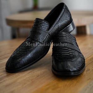 May include: A pair of black leather loafers with a snakeskin texture. The shoes are displayed on a wooden surface. The text "MenExoticLeather" is visible on the shoe.