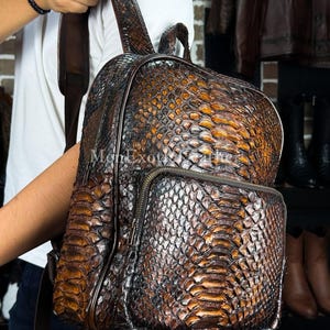May include: A brown leather backpack with a reptile skin pattern. The backpack has a front pocket with a zipper and adjustable straps. The bag is made of a dark brown leather with lighter brown and orange accents.