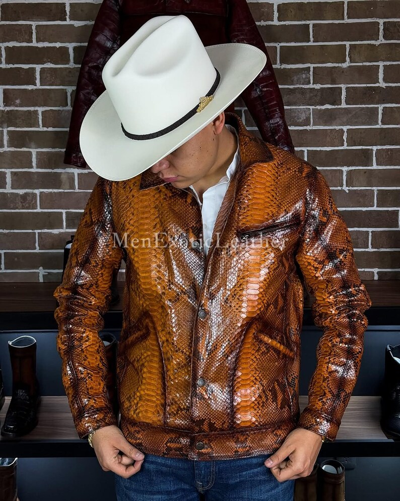 Op de afbeelding: Een bruine slangenleren jas met een knoopsluiting. De jas heeft een kraag en twee voorzakken. De persoon draagt een witte cowboyhoed met een zwarte band en gouden accent. De tekst "MenExoticLeather" is zichtbaar.