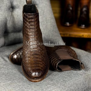 Puede incluir: Botas Chelsea de cuero marrón con un patrón texturizado similar al de un reptil. Las botas tienen una puntera redondeada y paneles laterales elásticos. La marca "Mon Exotic Leather" es visible.