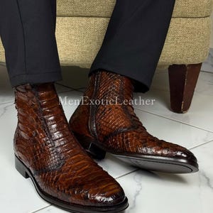 Op de afbeelding: Bruine leren enkellaarsjes met een reptielhuidtextuur. De laarzen hebben een zijrits en een lage hak. De laarzen worden gedragen met een zwarte broek. De tekst "MenExoticLeather" is zichtbaar.