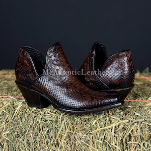 Könnte beinhalten: Braune Leder-Ankle Boots mit Schlangenmuster. Die Stiefel haben eine spitze Zehenpartie, einen kleinen Absatz und einen Seitenausschnitt. Sie werden auf Heu präsentiert, mit dem Text "Men exotic Leather" sichtbar.