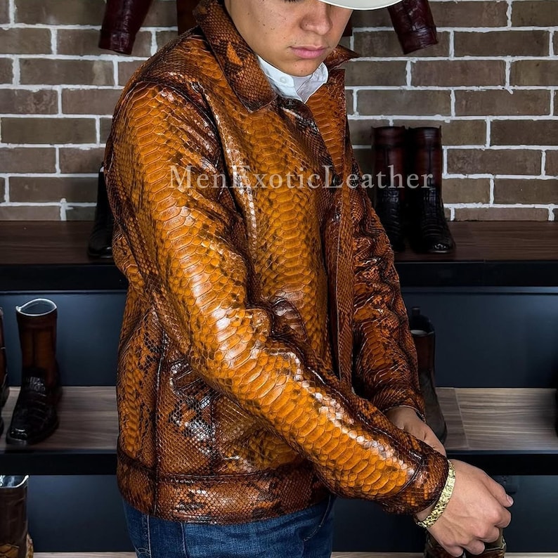 Op de afbeelding: Een bruin en tan slangenleerpatroon jasje met een kraag. De jas heeft een knoopsluiting aan de voorkant en lange mouwen. De tekst "Men Exotic Leather" is zichtbaar op de jas. De jas wordt gedragen met een blauwe jeans.