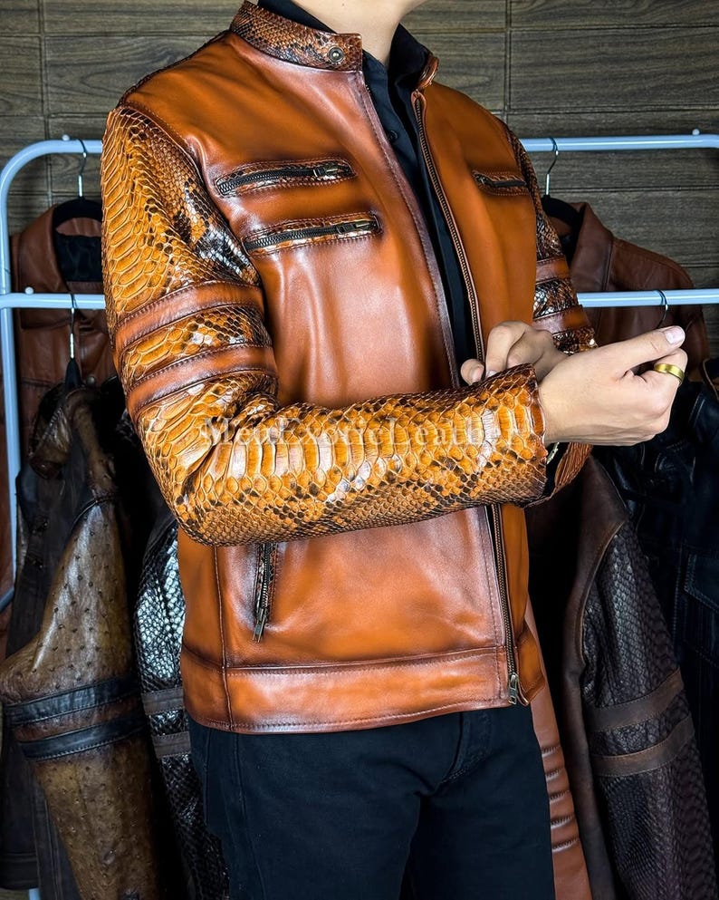 May include: A brown leather jacket with snake skin patterned sleeves and shoulder accents. The jacket has a zipper closure, multiple pockets, and a stand-up collar. Other leather jackets are visible in the background.