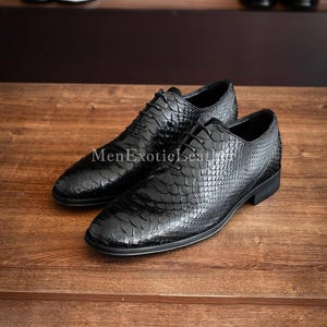 May include: A pair of black leather dress shoes with a textured, scale-like pattern. The shoes are laced up and sit on a wooden surface. The text "MenExoticLeather" is visible on the left shoe.