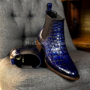 May include: Blue leather Chelsea boots with a crocodile skin pattern. The boots have a pointed toe and elastic side panels. The boots are on a grey upholstered chair. The text "MenExoticLeather" is visible.