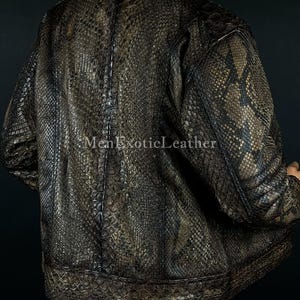 May include: A brown and black snakeskin-patterned leather jacket. The jacket has a bomber style with a ribbed hem and cuffs. The text "Men Exotic Leather" is visible on the bottom left of the image. The jacket is worn over blue jeans.