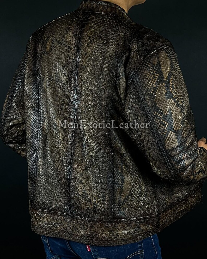 May include: A brown and black snakeskin-patterned leather jacket. The jacket has a bomber style with a ribbed hem and cuffs. The text "Men Exotic Leather" is visible on the bottom left of the image. The jacket is worn over blue jeans.