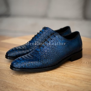 Op de afbeelding: Een paar blauwe leren geklede schoenen met een getextureerd, schubachtig patroon. De schoenen hebben zwarte veters en zolen, en de tekst "MenExoticLeather" is zichtbaar aan de zijkant. De schoenen worden gepresenteerd op een houten oppervlak.